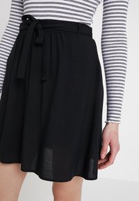 Kvinna klädd i en svart knälång kjol med knytbälte och en långärmad vit topp med tunna svarta horisontella ränder.