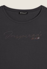 Camiseta gris oscuro con escote redondo. Presenta la palabra "Inspired" en pedrería y "HEART" en letras metálicas y mayúsculas de estilo bloque. Textura de tela suave.