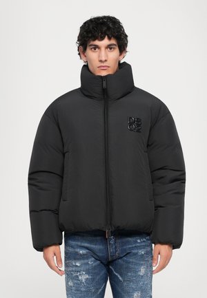 Junger Mann, der eine schwarze Steppjacke mit hochgestelltem Kragen und DSQ2-Logo auf der Brust trägt, kombiniert mit abgenutzten blauen Jeans, stehend vor einem weißen Hintergrund.