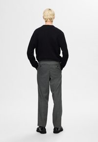 Schwarzer Strickpullover und graue gemusterte Hose mit flachen Gesäßtaschen, getragen mit schwarzen Schuhen. Von hinten angezeigt.