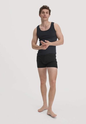 Schwarzes Tanktop und schwarze Boxershorts an einer stehenden männlichen Figur mit heller Haut und kurzen, welligen Haaren, barfuß vor einem schlichten Hintergrund.