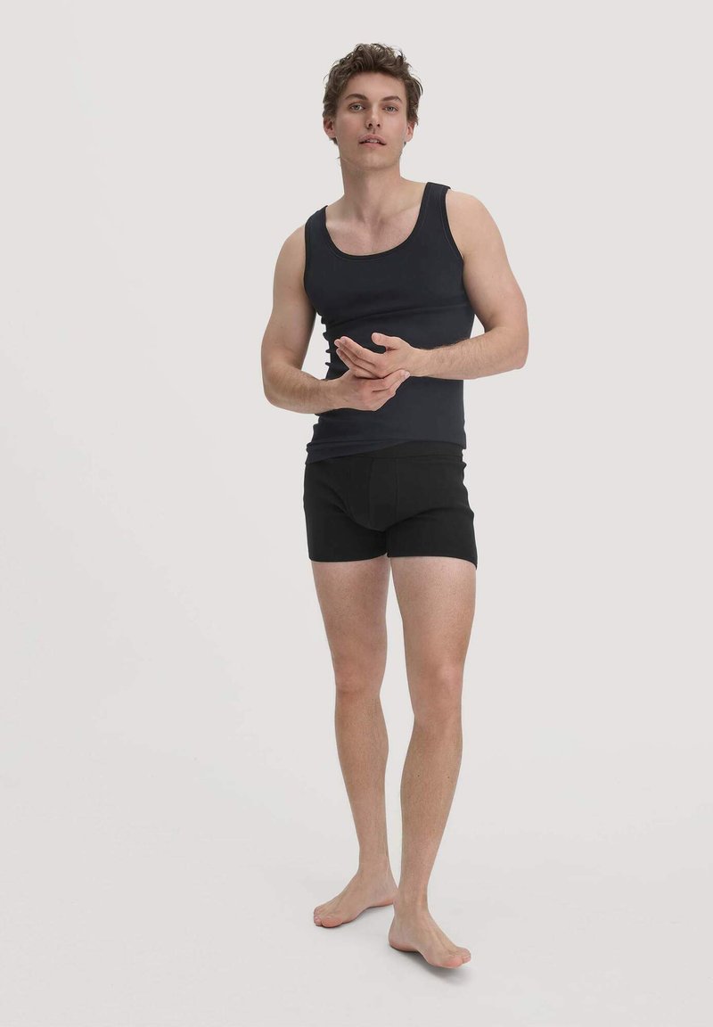 Zwarte tanktop en zwarte boxershorts op een staande mannelijke figuur met een lichte huid en kort, golvend haar, blootsvoets tegen een effen achtergrond.