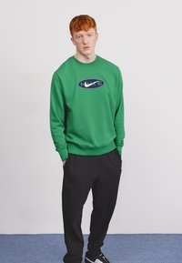 Sweatshirt verde com logotipo frontal; design de gola redonda; combinado com calças pretas; corte descontraído; textura suave; usado com ténis pretos.