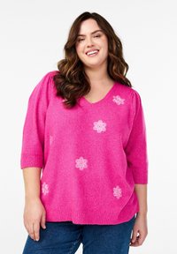 Rosa Pullover mit kurzen Ärmeln, V-Ausschnitt und weißer floralem Stickerei. Weicher Stoff und lässige Passform. Getragen mit blauen Jeans.