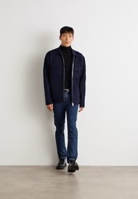 Mornarsko zadrgo zaprt jopič z dvema sprednjima žepoma, nošen čez črn rolak, v kombinaciji z modrimi kavbojkami in črnimi čevlji. Gladka tekstura, sodoben kroj.