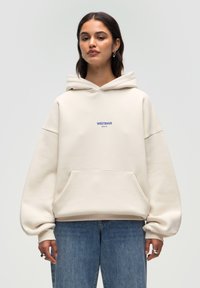 Beige übergroßer Kapuzenpullover mit einer Vorderseite Tasche, bedruckt mit "WRSTBHV" in blauer Schrift. Der Stoff ist weich mit einer strukturierten Oberfläche.