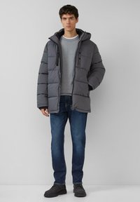 Grijze donsjack met capuchon, met een horizontaal doorgestikt ontwerp, zwarte accenten en knopen aan de voorkant. Gecombineerd met een grijs gestreept shirt en blauwe spijkerbroek.