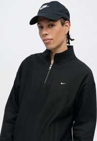 Fekete cipzáras kapucnis pulóver magas gallérral és fehér Nike logóval a mellrészen. Fekete sapkával párosítva, amelyen fehér logó található.