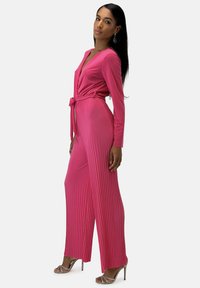 Una tuta rosa a maniche lunghe con scollatura a V profonda e texture plissettata, legata in vita. Abbinata a tacchi a spillo argento con cinturini.