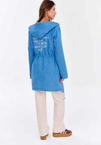 Kobieta z brązowymi włosami ubrana w niebieską kurtkę z kapturem z białym napisem na plecach, beżowe spodnie i brązowe sneakersy na platformie, stoi w pozycji bocznej.