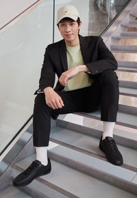 Černý blejzr na světle zeleném tričku, světlá kšiltovka, černé tenisky, pohodlně sedící na šedých schodech s proskleným zábradlím v moderním prostředí.