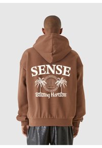 Haut à capuche marron avec un grand motif blanc représentant des palmiers et le texte "SENSE" et "Blazing Horizon" au dos. Tissu doux, coupe oversize.