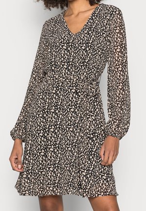 Woman wearing a black and beige spotted wrap dress with long sheer sleeves and a V-neckline, tied at the waist with a fabric belt.