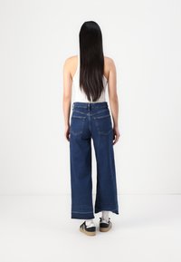 Pantalones de mezclilla de pierna ancha con un lavado azul oscuro, dobladillo sin terminar y dos bolsillos traseros, combinados con una camiseta sin mangas blanca y zapatillas negras.