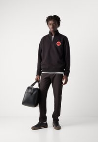 Sudadera negra con cremallera y un logo rojo, combinada con pantalones marrones oscuros y zapatillas negras. El modelo sostiene un bolso de cuero negro.