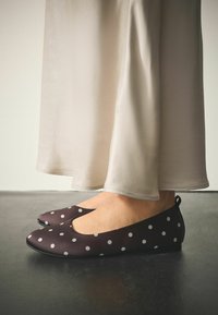 Braune Ballerinas mit Polka-Dot-Muster, glatter Textur, abgerundeter Spitze und niedrigem Profil, kombiniert mit einem fließenden cremefarbenen Kleid.