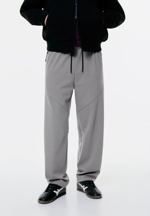 Hellgraue Sweatpants mit lockerer Passform, gesteppten Nähten, elastischem Bund und seitlichen Taschen. Getragen mit schwarzen Turnschuhen.