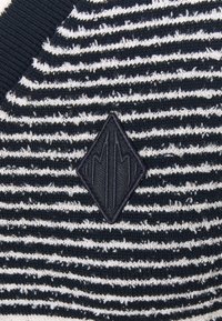 Tessuto a maglia a strisce bianche e nere con un emblema ricamato a forma di diamante nero che presenta dettagli astratti a linee verticali.