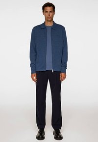 Blå jacka med dragkedja i texturerat tyg, kombinerad med en ljusblå skjorta och mörka byxor. Har en krage och en minimalistisk design.