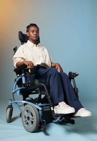 Ηλεκτρικό αναπηρικό αμαξίδιο με μαύρη έδρα, μπλε σκελετό και μεγάλα γκρι τροχούς. Ο χρήστης φοράει ένα ριγέ μπεζ πουκάμισο και σκούρο παντελόνι με λευκά αθλητικά παπούτσια.