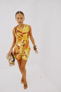 Robe-culotte jaune à motifs avec des accents marron et vert, dotée d'un design sans manches et de détails en boutons, associée à une pochette en perles et des sandales.