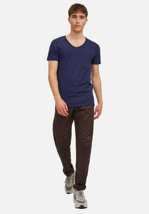 Young man walking forward wearing a navy blue V-neck T-shirt, dark brown pants, and beige athletic shoes on a white background.