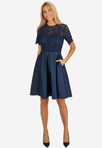 Navyblaues Kleid mit einem Spitzenoberteil mit filigranen Ausschnitten und kurzen Ärmeln, ausgestelltem satinierten Rock mit Taschen und einer glatten Textur.