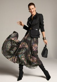 Schwarze taillierte Blazer mit einem Rosenakzent, kombiniert mit einem gefalteten, gemusterten Maxirock in dunklen Farbtönen und bunten Designs. Schwarze Stiefeletten und eine Handtasche.