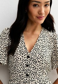 Kurzes, ärmelloses Kleid aus einem beigen Stoff mit schwarzem Punkte-Muster, V-Ausschnitt und schwarzen Knopfakzenten. Strukturiert Oberfläche mit gerafftem Taillenbereich.