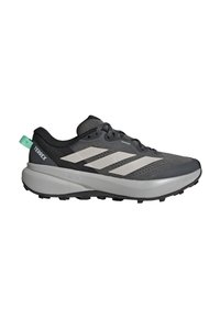 Μαύρο και γκρι παπούτσι τρεξίματος Adidas Terrex trail με λευκές ρίγες, χοντρή μαλακή σόλα και στρογγυλό πράσινο λουράκι στη φτέρνα σε λευκό φόντο.
