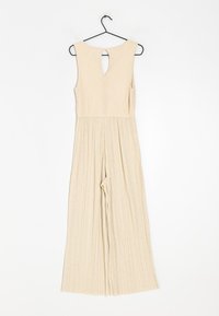 Monos con un diseño sin mangas, de tela texturizada y piernas anchas con pliegues, en color beige y con un escote en la espalda. Cuela de un gancho negro.