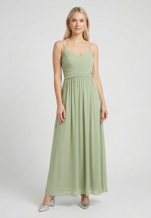 Frau trägt ein bodenlanges, hellgrünes Spaghetti-Trägerkleid mit gerafftem Mieder und silbernen Absätzen, steht vor einem einfarbigen Hintergrund.