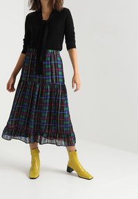 Svart långärmad topp med knytdetalj, tillsammans med en rutmönstrad, lager-på-lager kjol i svart, grönt och rött. Gula klackboots som slutar vid ankeln fulländar outfiten.