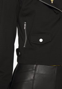 Zwarte cropped jacket met een ritszak en drukknoopdetails. Heeft een geweven stof en een zilveren ritsaccent. Contrastvoering zichtbaar bij de zoom.