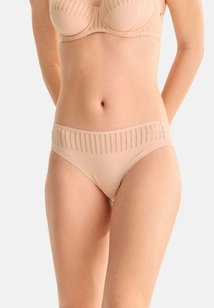 Femme portant un soutien-gorge et une culotte beige assortis avec des détails de rayures verticales transparentes sur la taille et les bonnets du soutien-gorge sur un fond uni.