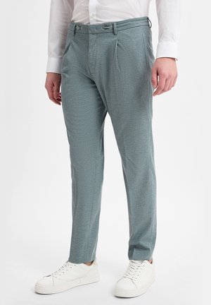 Mann trägt hellgraue, maßgeschneiderte Hose, weißes Hemd und weiße Sneakers, steht vor weißem Hintergrund.