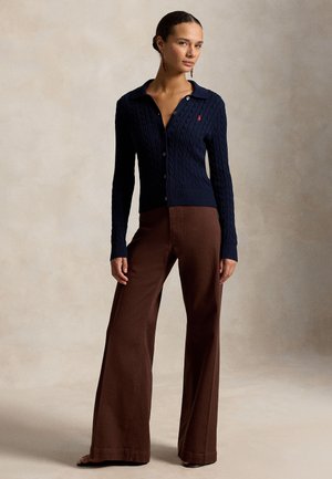 Žena stojaca v námornícky modrom kardigane s vrkočovým vzorom a hnedých širokých nohaviciach na jednoliatom béžovom pozadí.