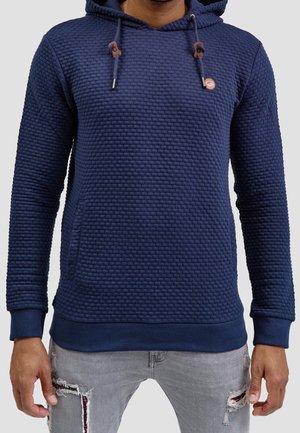 Homme portant un sweat à capuche bleu marine texturé avec cordons et un jean gris clair déchiré, debout les bras détendus le long du corps.