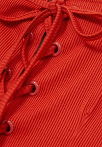 Tissu rouge côtelé avec un design à lacets et un nœud sur le dessus. Comprend des œillets rouges brillants et une texture lisse.
