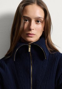 Pull en tricot bleu marine avec un col haut et des détails de fermeture éclair dorés, présentant un motif texturé et une coupe décontractée.