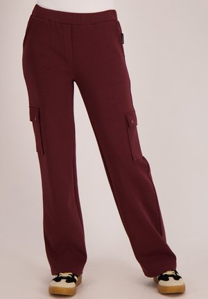 Maroon cargobroek met zijflapzakken, gedragen met zwart-witte sneakers op een effen achtergrond.