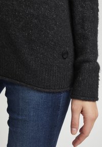 Pull en tricot gris foncé avec une finition texturée, ourlet côtelé et un bouton noir sur le côté gauche. Associé à un jean bleu foncé.