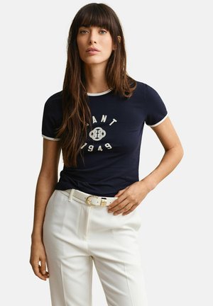 Vrouw met lang bruin haar, gekleed in een marineblauw T-shirt met logo en witte broek met hoge taille en witte riem, staand tegen een witte achtergrond.