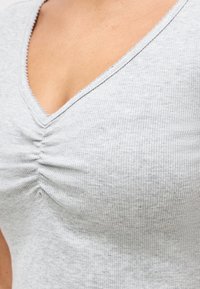 Κοντινό πλάνο ανοιχτόγκρι ριγωτού τοπ με λαιμόκοψη σε σχήμα V και λεπτομέρεια μαζεμένου υφάσματος στο κέντρο μπροστά.