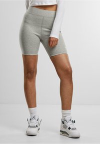 Donna che indossa pantaloni biker grigi aderenti, un crop top bianco a maniche lunghe, calze bianche e sneakers alte bianche, in piedi su un pavimento bianco.
