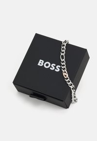 Pulsera de cadena de metal plateado con secciones interconectadas, con un cierre de acento en oro rosa, situada sobre una caja rectangular negra etiquetada como "BOSS".