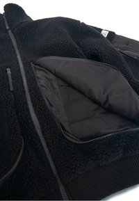 Czarna polarowa kurtka z zamkiem błyskawicznym, o miękkiej teksturze, z boczną kieszenią i gładką podszewką. Detalem jest ściągacz na dole.