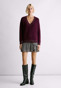 Maglione a V in maglia bordeaux con orlo texturizzato abbinato a una mini gonna a volant in bianco e nero e stivali neri in pelle alti fino al ginocchio.