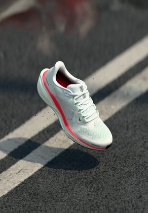 White athletic shoe with a pink accent, suspended mid-air over a textured asphalt surface with faint tire markings.