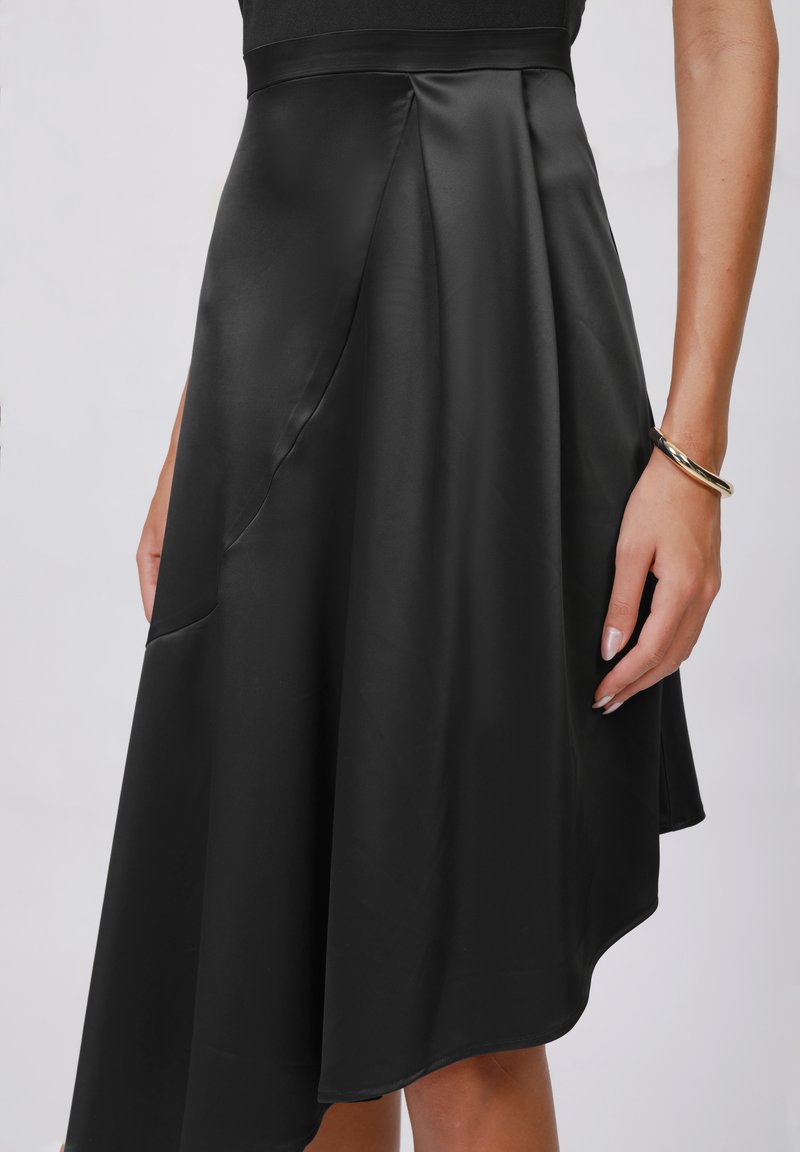 Zwarte satijnen midi-rok met een asymmetrische zoom, voorzien van een zachte textuur, plooien en zijzakken, gecombineerd met een gouden armband.